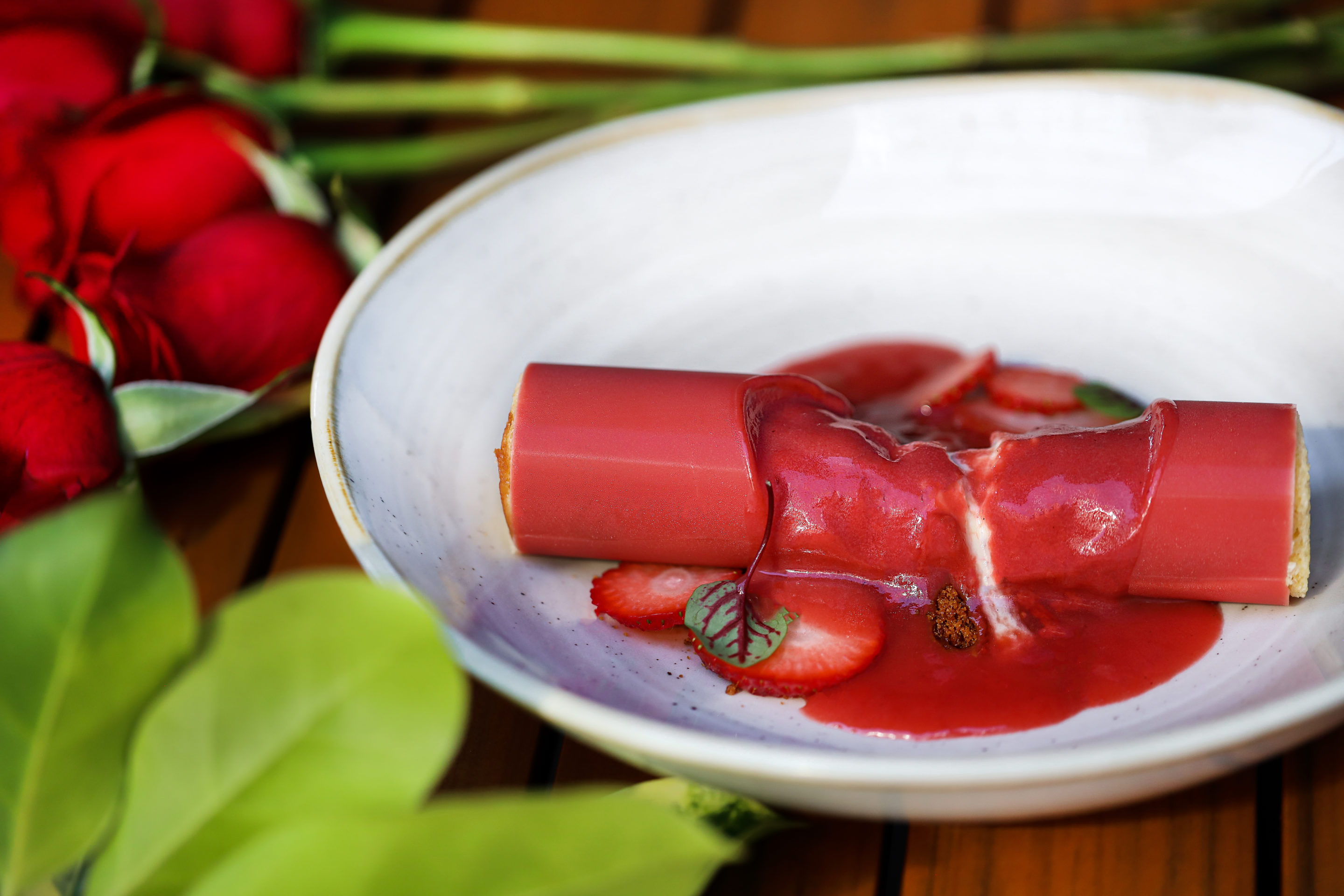 red log-shaped dessert in white dish with roses in the background