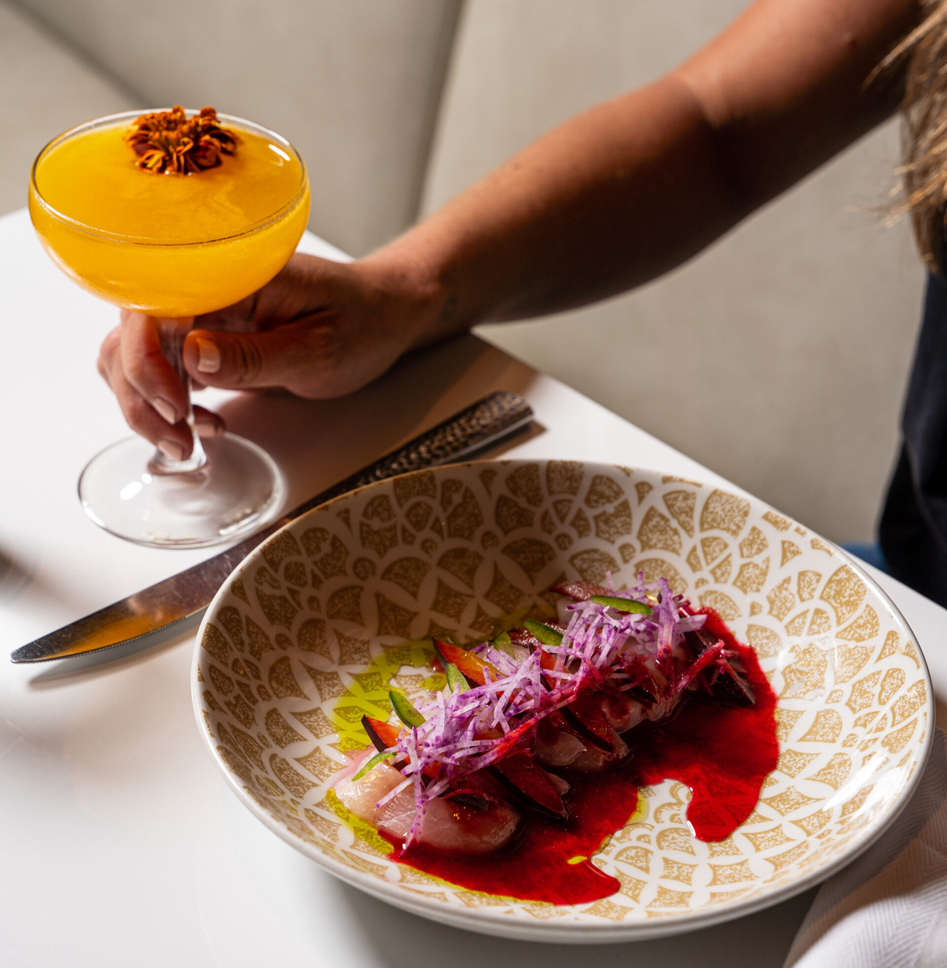 Colorful plated dish with an orange cocktail plus a garnish being held next to it.
