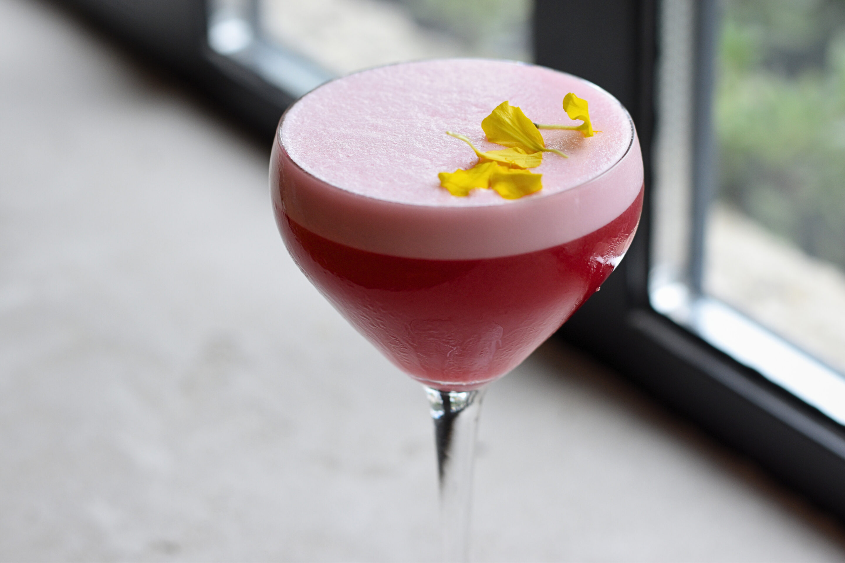 Pink cocktail in a glass with a yellow flower garnish.
